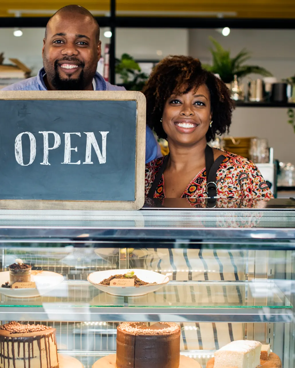 Two bakery owners behind their cake display, holding an OPEN sign